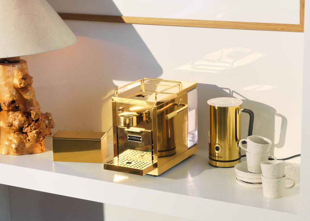 Sjöstrand kaffemaskin och mjölkskummare i guld på en vit hylla med kaffekoppar